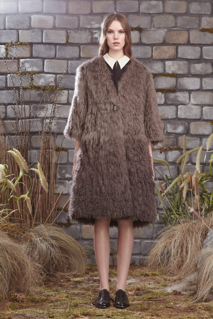 Woman in a fluffy brown coat, white-collared shirt, and black shoes, standing by a brick wall with dry plants.