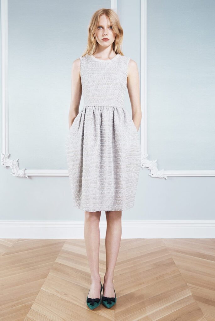 Woman in a sleeveless light gray textured dress standing on a wooden floor with hands in pockets.