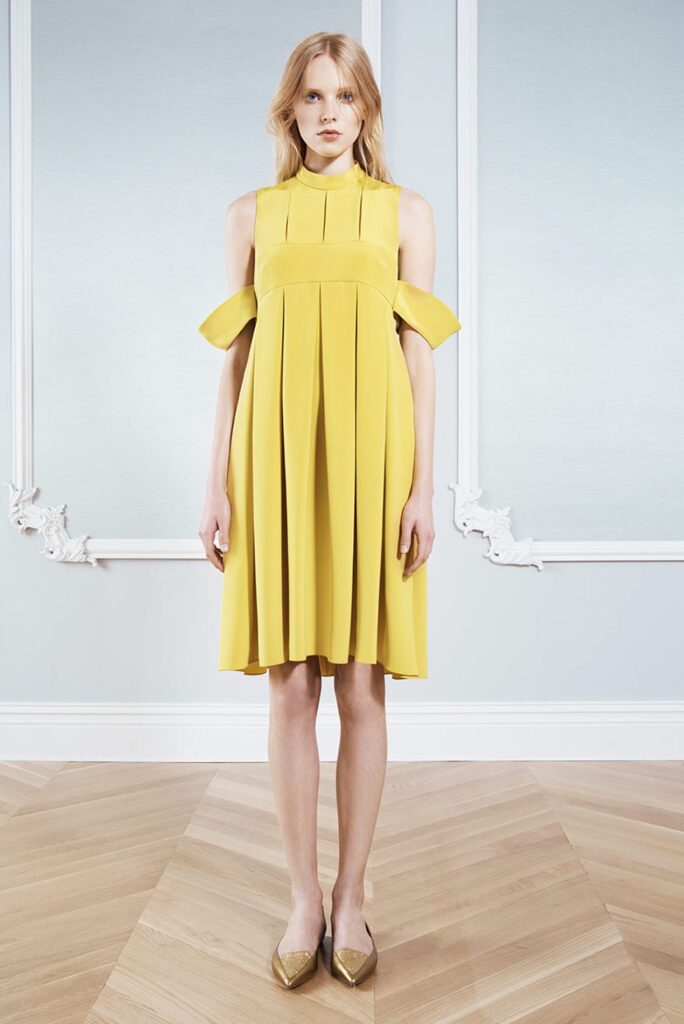 Woman stands in a yellow pleated dress with unique cut-out sleeves, on a wooden floor, against a pale wall.