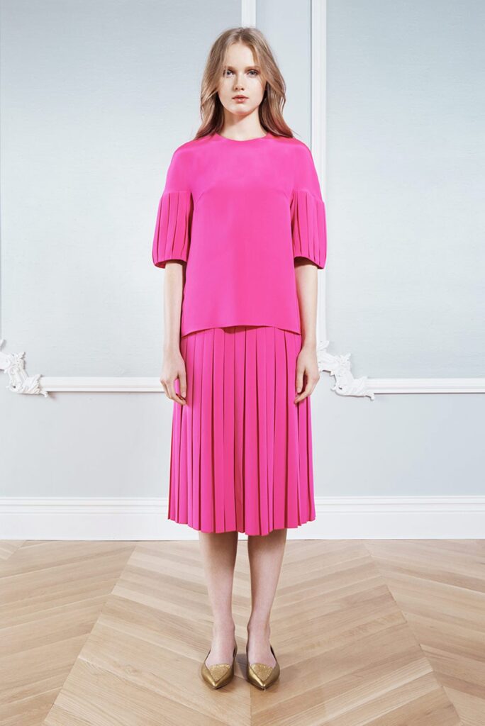 Woman in a bright pink pleated outfit stands indoors on a wooden floor, wearing gold pointed shoes.