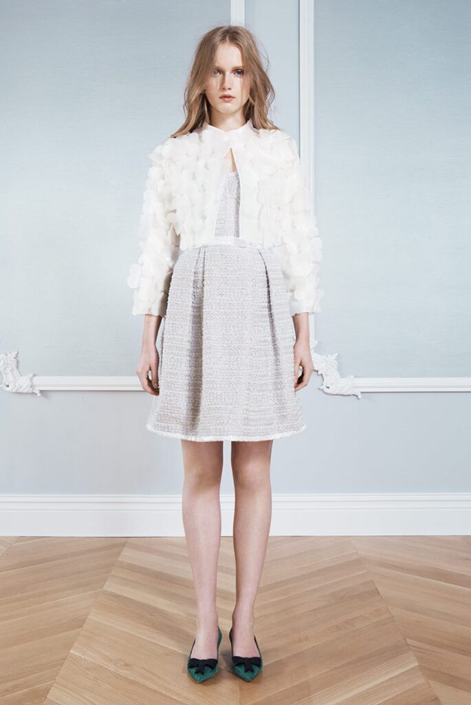 Woman in a white textured dress and floral jacket standing on a wooden floor in a minimalist room.