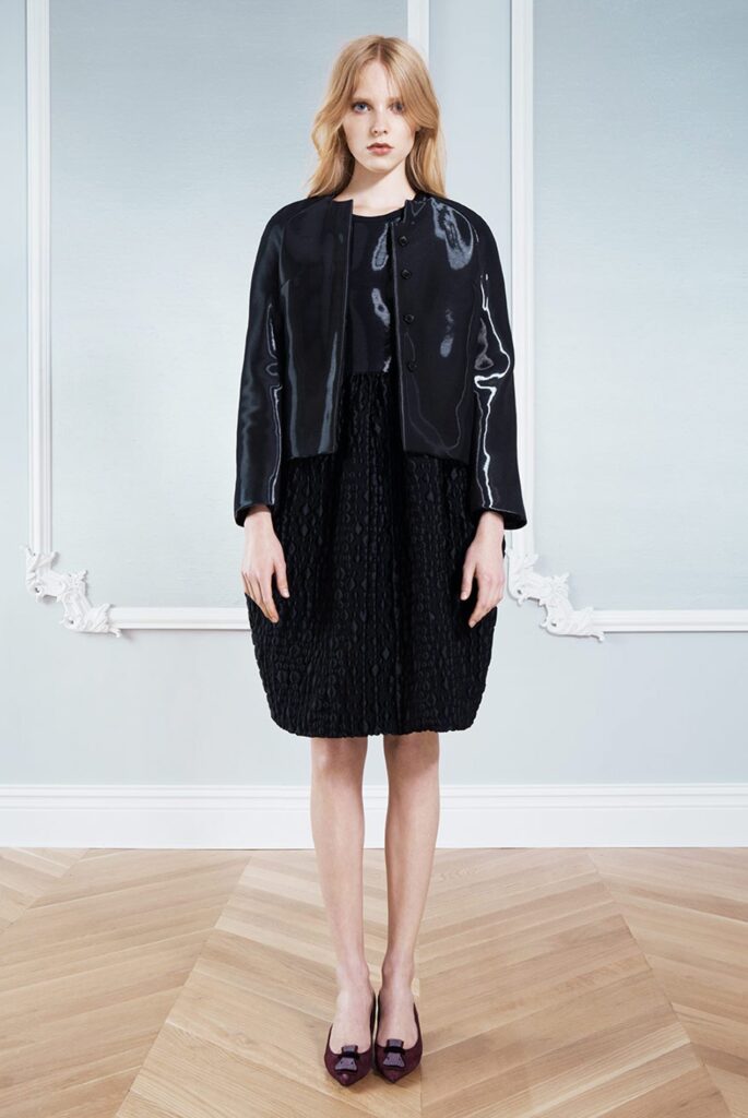 Woman in a black textured dress, shiny black jacket, and purple shoes stands in a light room.