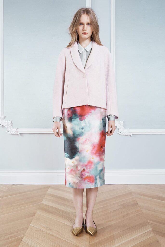 Woman in a pale pink blazer, gray blouse, and colorful watercolor skirt, standing indoors on wood floor.