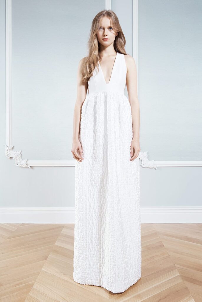 Woman in a sleeveless white gown with a deep V-neck, standing on wooden floor against a pale blue wall.