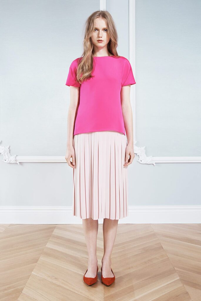 Woman in a bright pink top and white pleated skirt stands indoors on a wooden floor, facing forward.