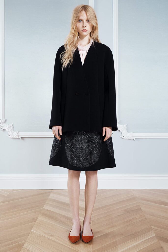 Woman in a black coat and skirt, pink blouse, and brown shoes stands on a wooden floor by a light wall.