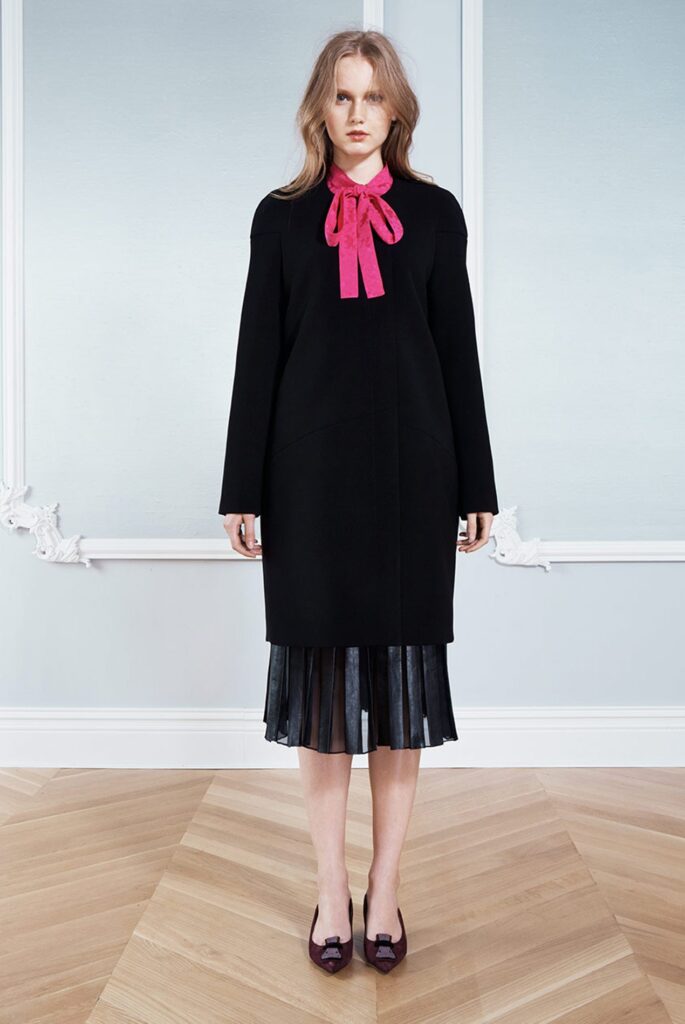 Woman in black dress with pleated skirt, pink bow at collar, standing on wooden floor in a light room.