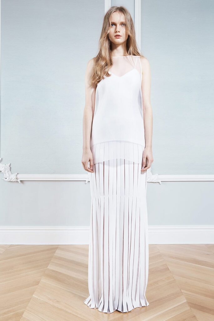 Woman in a sleeveless white top and long pleated white skirt stands in a minimal, light blue room.