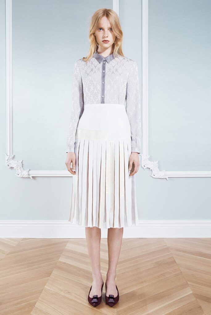 Woman in a light gray lace blouse and white pleated skirt, standing on a wooden floor in a pale room.
