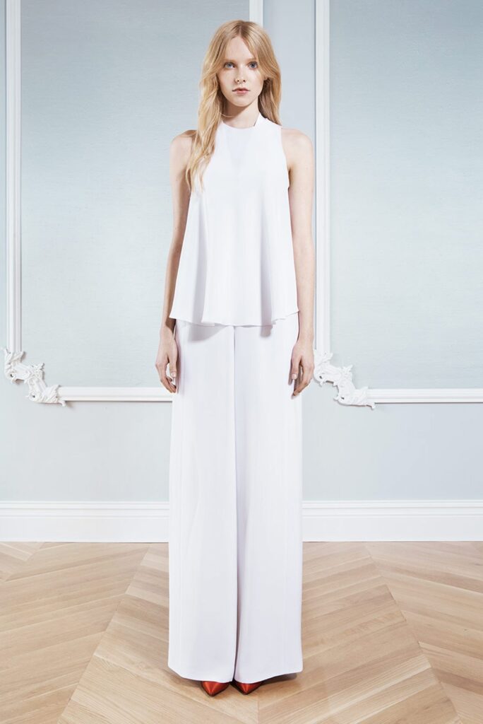 Woman in sleeveless white top and wide-leg pants stands on wood floor against a light blue wall.