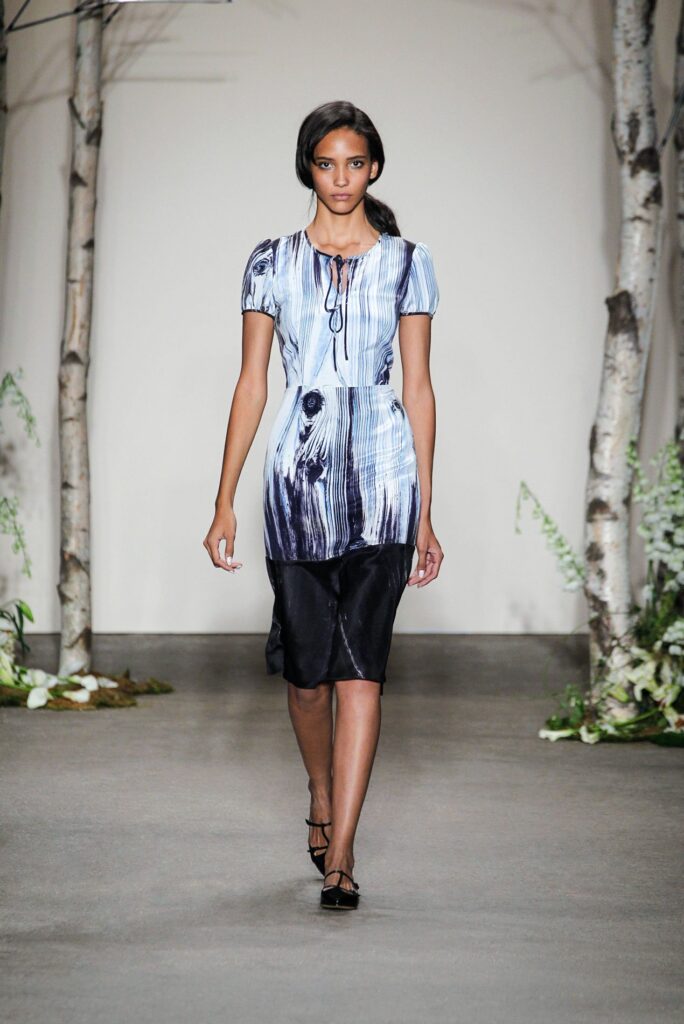 Model walks runway in blue and black dress with tree pattern, flanked by birch trees and greenery.