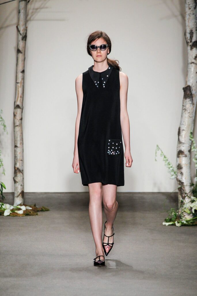Woman on runway in sleeveless black dress, black sunglasses, and pointed shoes, with trees in background.