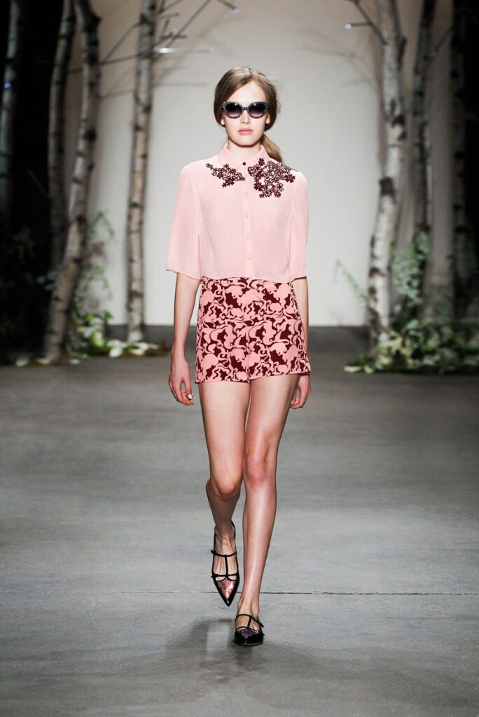 Model in pink blouse with jeweled collar and printed shorts walks runway; birch trees in background.