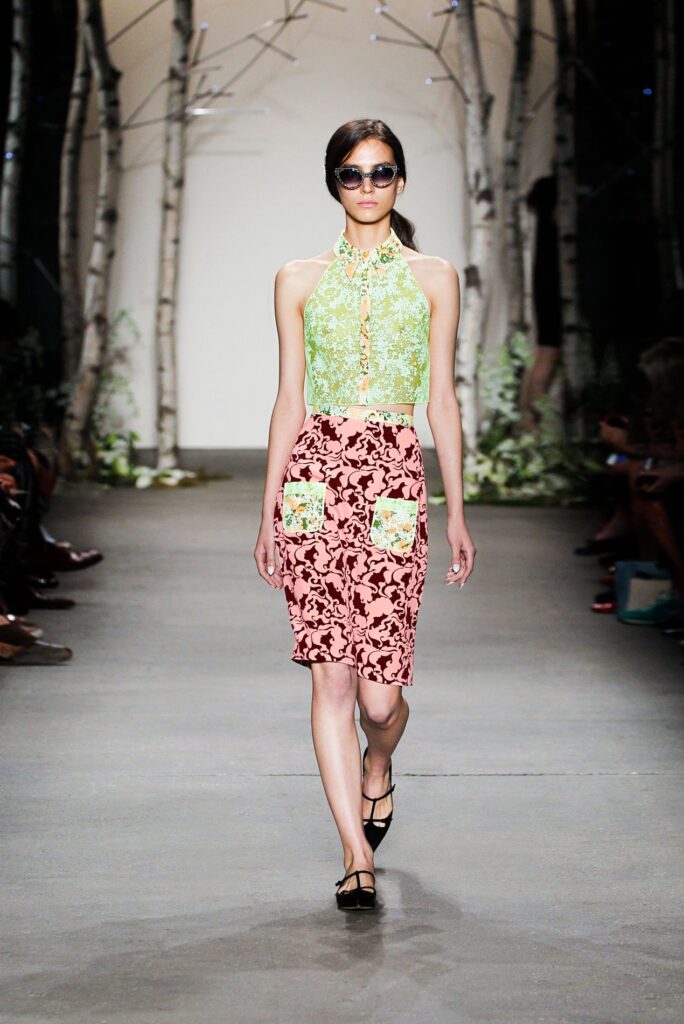 A model walks a runway in a green lace top and pink floral skirt with pockets, wearing sunglasses.