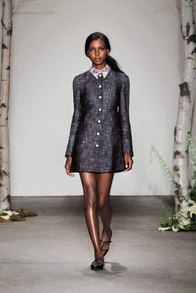 A model walks on a runway in a gray long-sleeve dress with white buttons and a beaded collar.