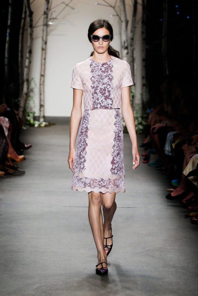 Model in sunglasses wearing a short-sleeve, knee-length pink lace dress walks on a runway with birch trees.