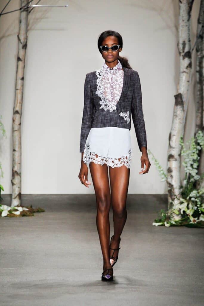 Model walks runway in a lace-trimmed jacket and skirt, wearing sunglasses, with tree props in background.