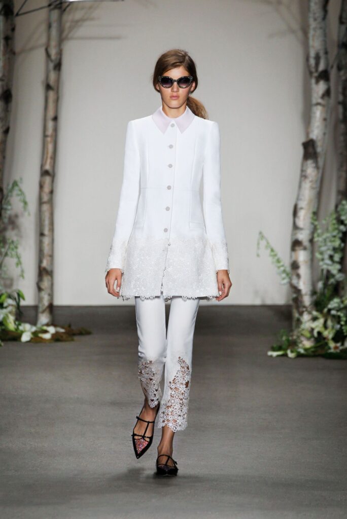 Model in white jacket and lace pants walks runway, wearing sunglasses; trees and plants in background.