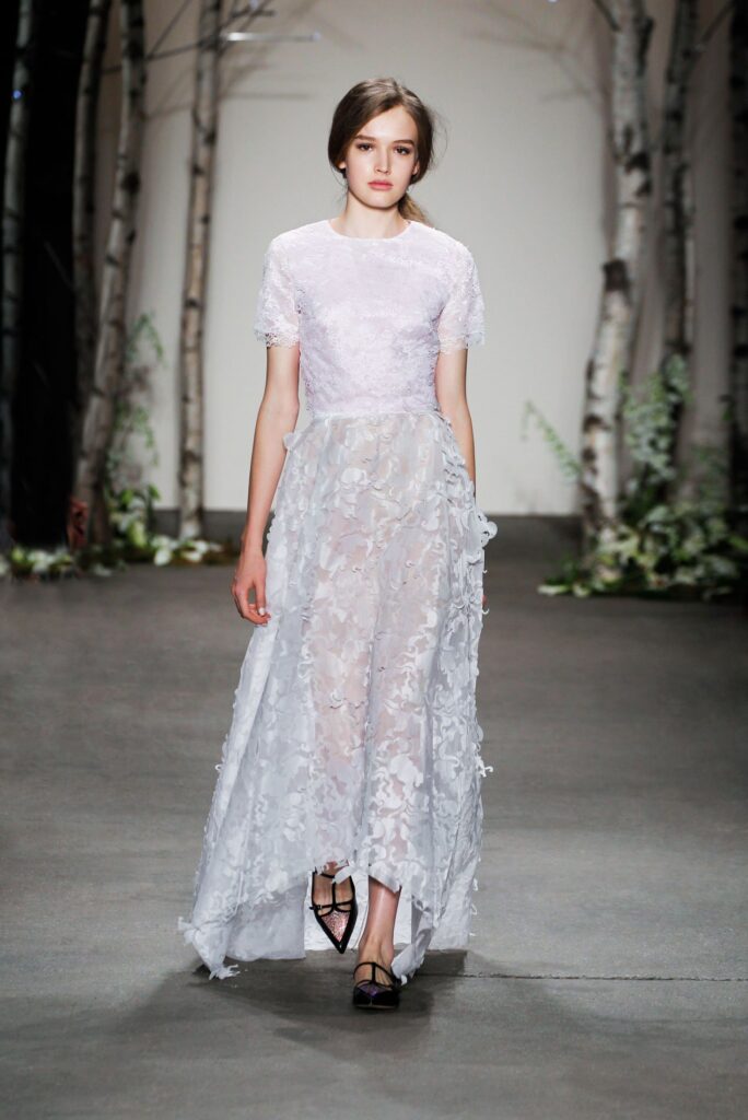 A model walks a runway in a sheer, white floral dress with short sleeves and flat black shoes.