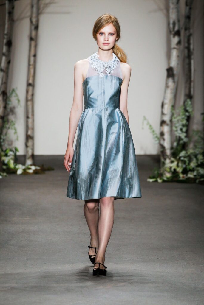 A model walks a runway in a shiny blue dress with a white lace collar, surrounded by birch trees.