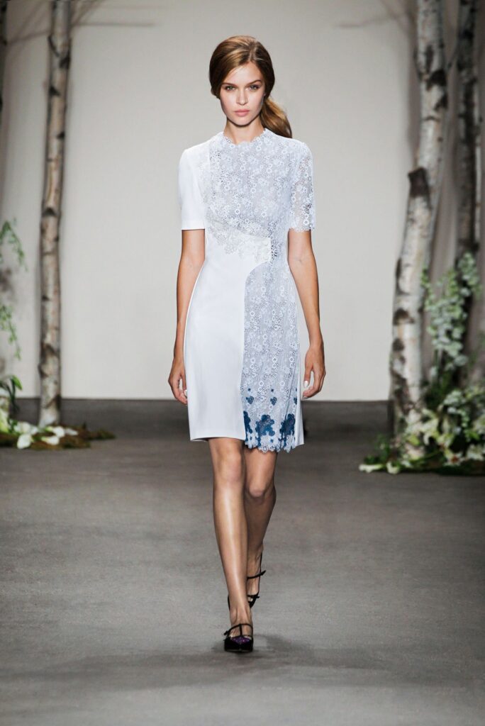 A woman in a white lace dress with blue accents walks on a runway with birch trees and greenery.