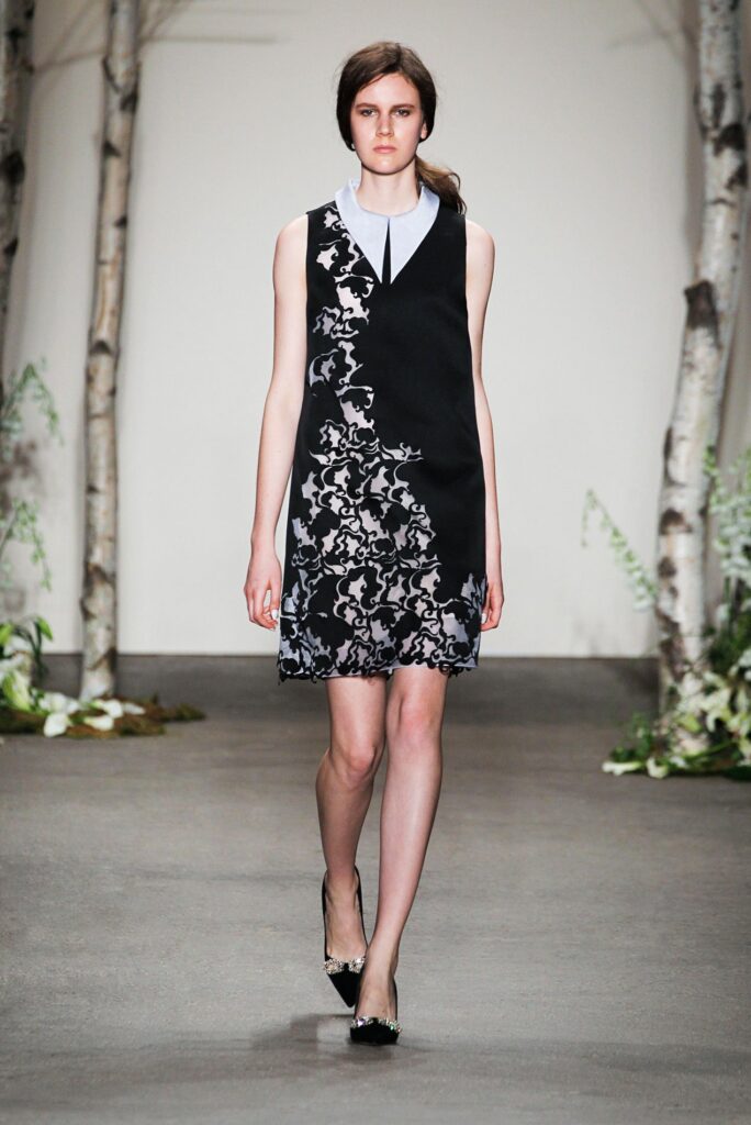 A woman models a sleeveless black dress with white floral patterns, walking on a runway with birch trees.