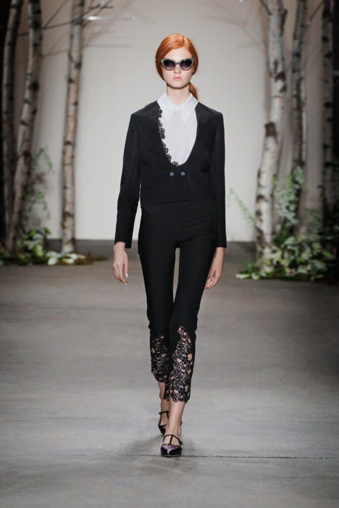 Model walks runway in black suit with lace pants, white shirt, sunglasses, and open-toe heels; forest backdrop.