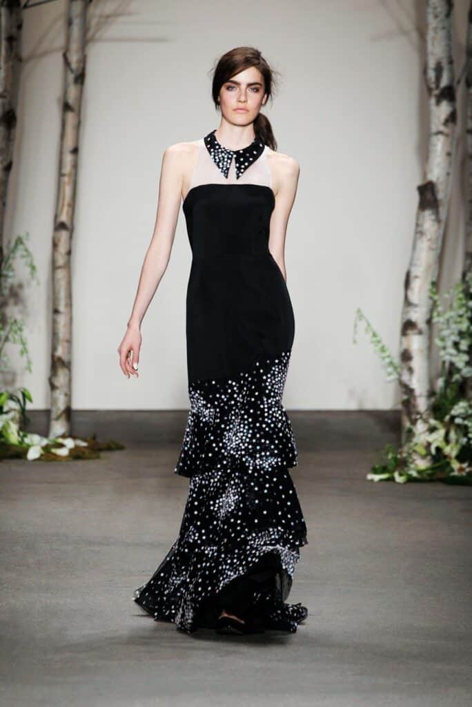 Model in black halter gown with white polka dots, ruffled skirt, walking on runway with birch trees.