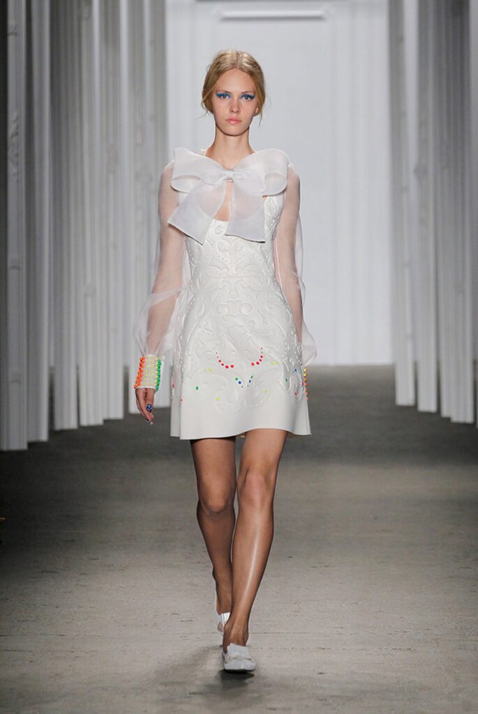 Model in a white dress with sheer sleeves, a large bow, and colorful accents walks a runway indoors.