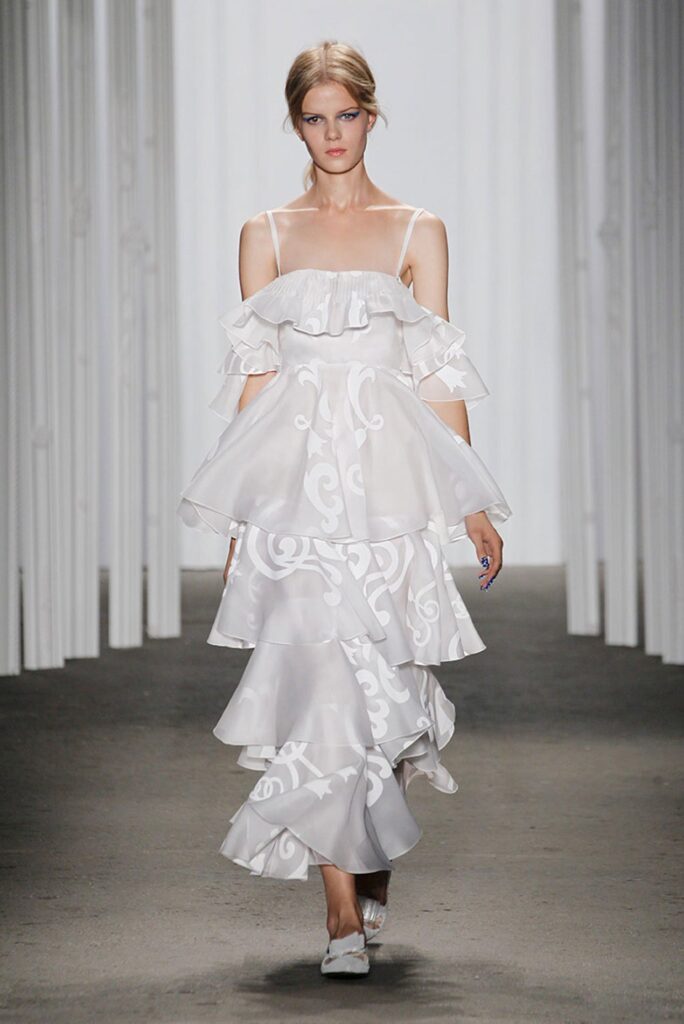 Woman in a layered white ruffled dress with thin straps walking on a runway in a minimalist setting.
