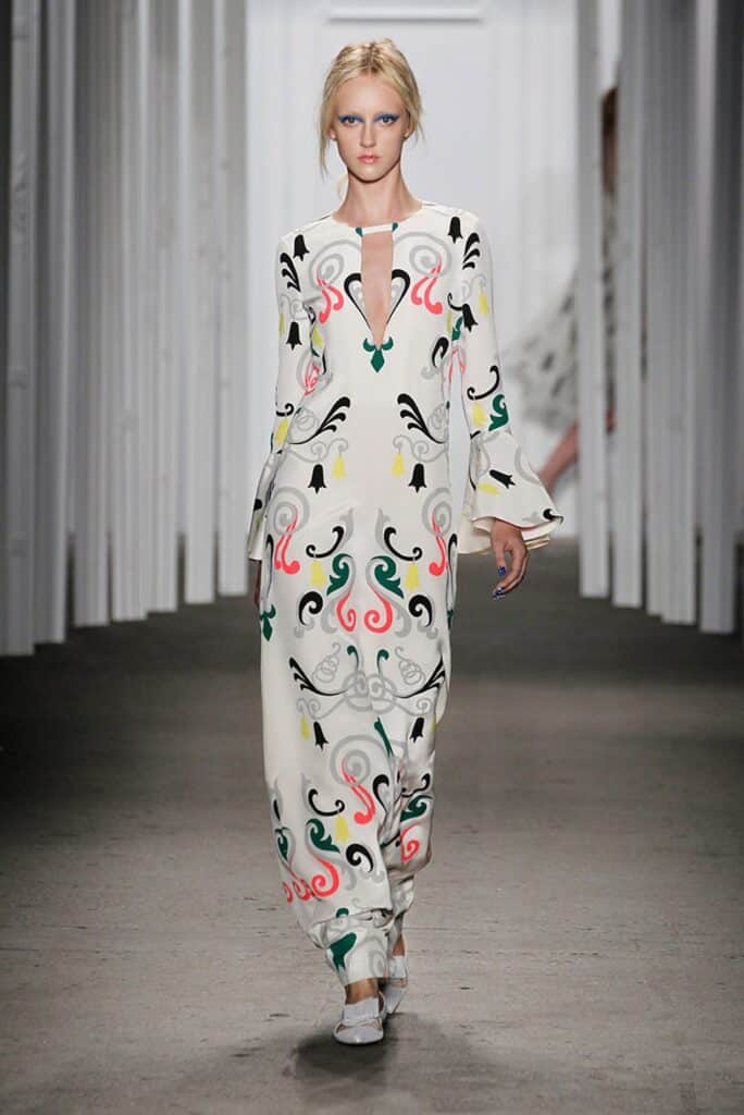 Model in a white jumpsuit with colorful abstract patterns walks down a runway in a minimalist setting.