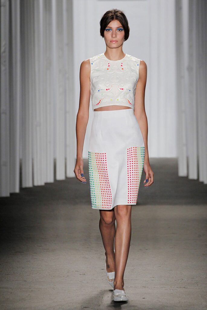 Model in a white crop top and skirt with colorful square patterns, walking on a runway.