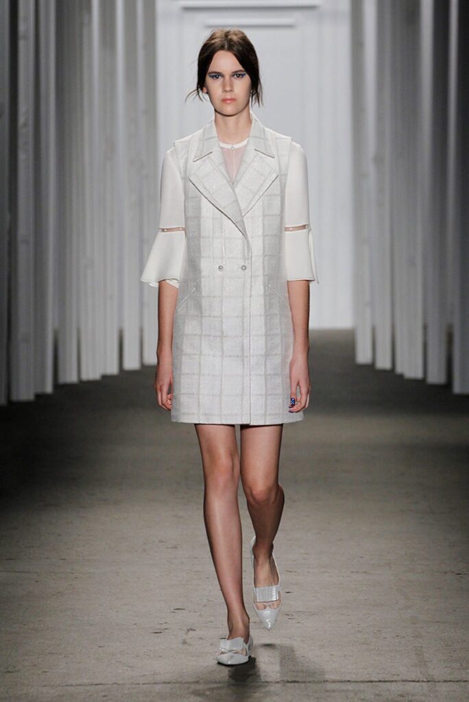 Model walks a runway wearing a white dress with sheer sleeves and white shoes in a modern, minimal setting.