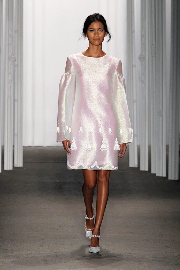 Model walks runway in a shiny, pale pink dress with tassels and puffed sleeves, wearing silver heels.