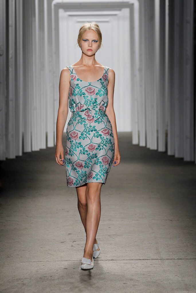 Model walks runway in a sleeveless, floral-patterned dress with a fitted waist, in a modern white hallway.