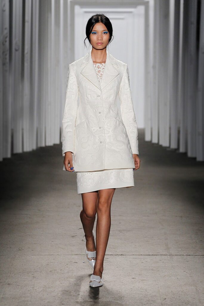 Model walks on runway wearing a white embroidered jacket and skirt set with white shoes, in a bright hallway.