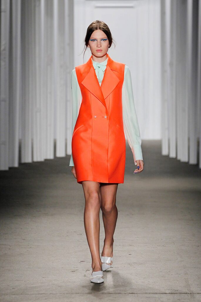 Model walks runway in bright orange sleeveless dress over a pale blue blouse and white heels.