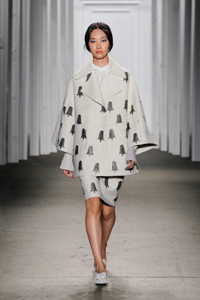 A model walks a runway in a white outfit with black abstract prints, featuring a cape and skirt.