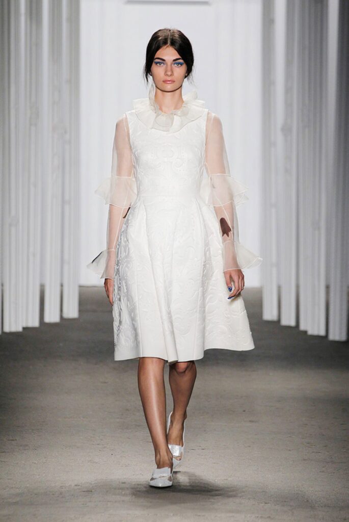 Model in a white, knee-length dress with sheer sleeves walks down a runway with a neutral background.
