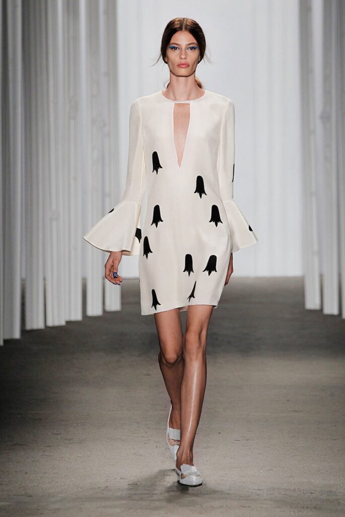 A model walks a runway in a white dress with black abstract shapes and flared sleeves.