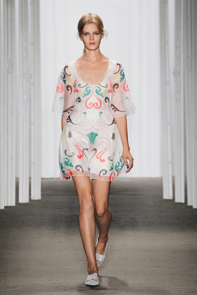 Model walks runway in a sheer white dress with colorful swirl patterns and white flats.