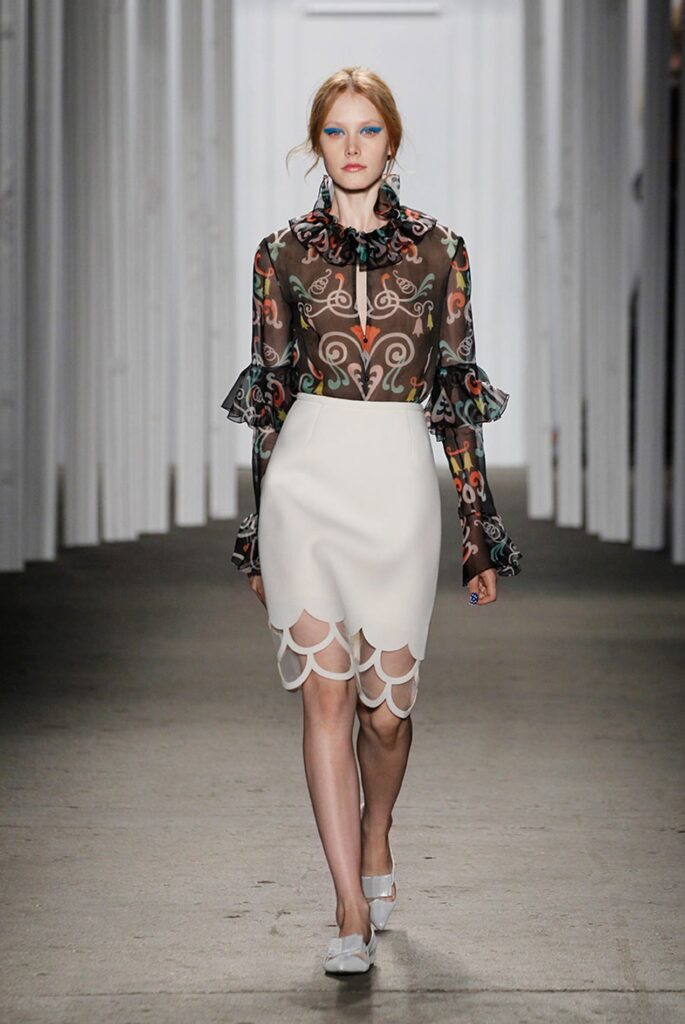 Model in a sheer, patterned blouse and white scalloped skirt walks down a fashion runway.