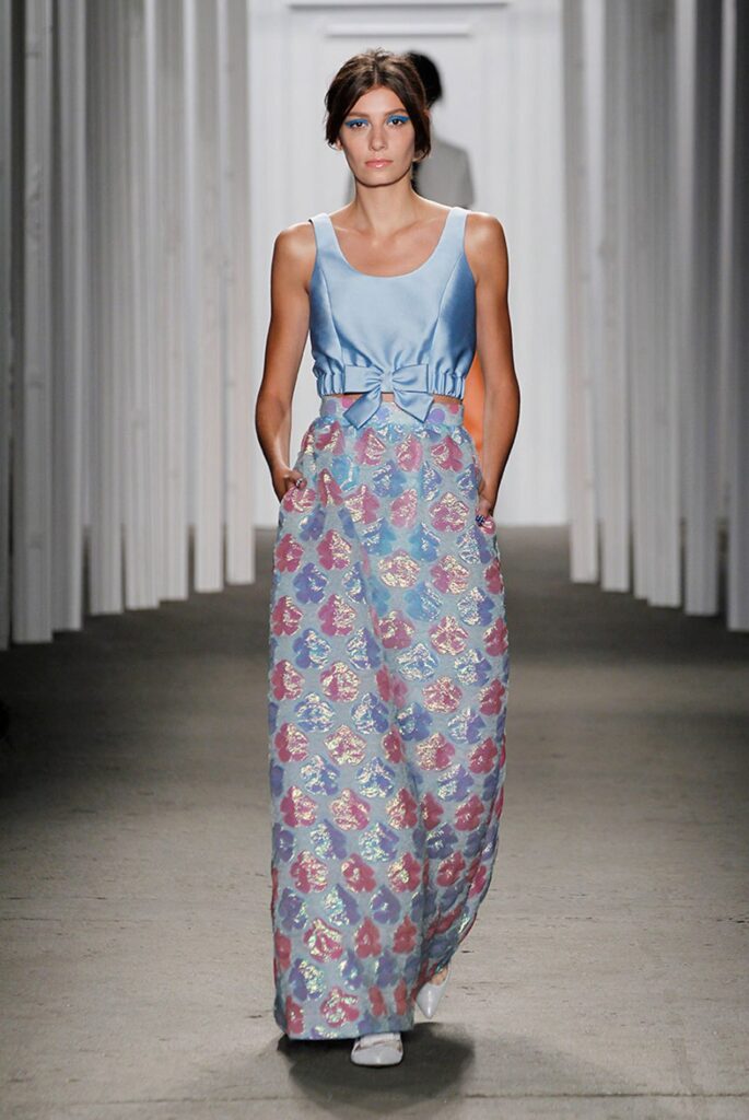 Model in a sleeveless blue top and a long, shimmery floral skirt walks down a runway.