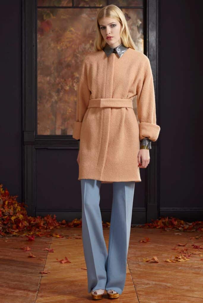 Woman in peach belted coat, blue pants, and gold shoes stands indoors with autumn leaves on the floor.