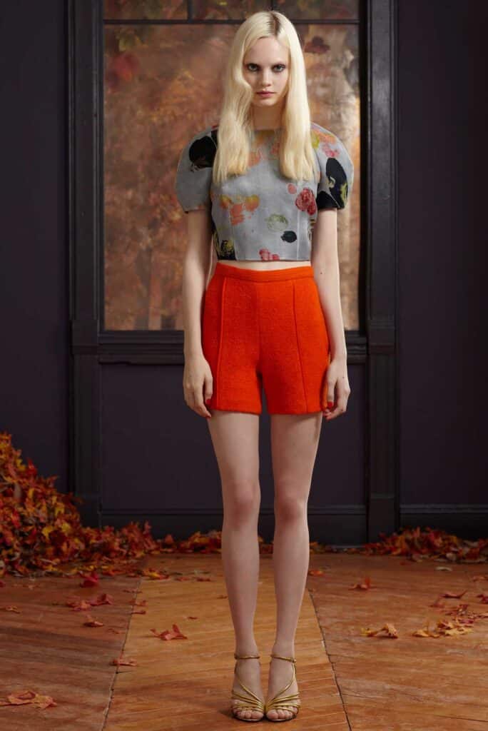 Woman in floral crop top and red shorts stands indoors on a wooden floor with autumn leaves around her.