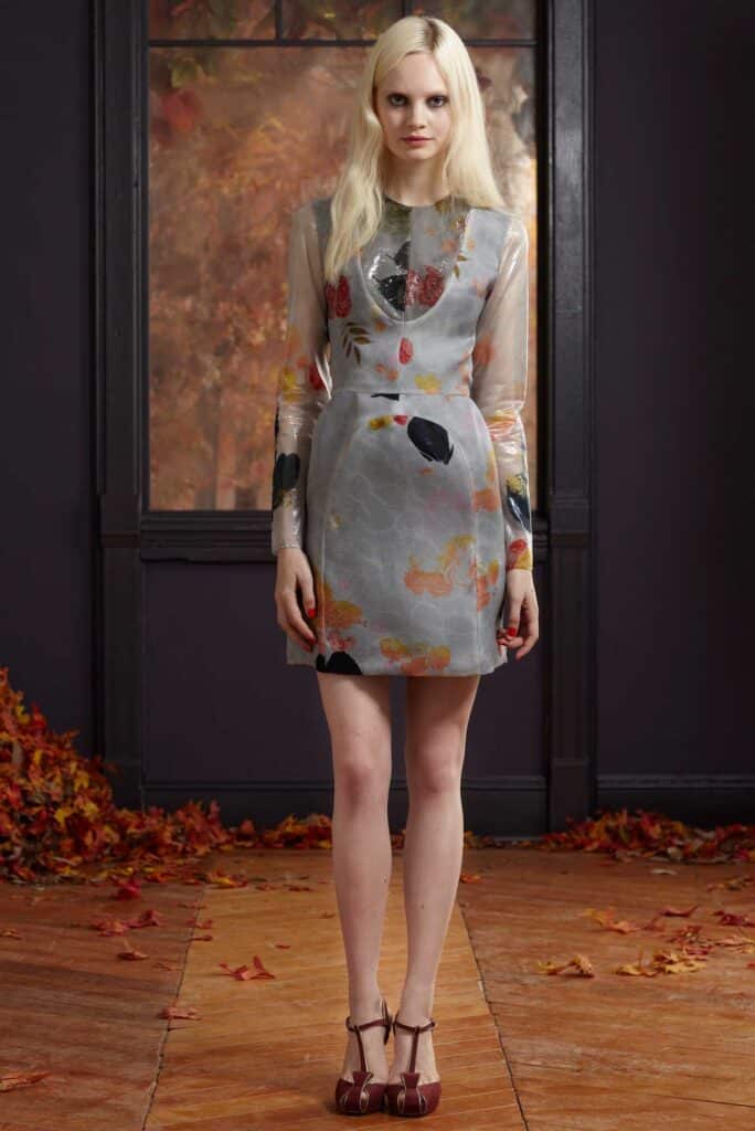 Woman in a gray floral dress and red shoes stands indoors on a floor covered with autumn leaves.