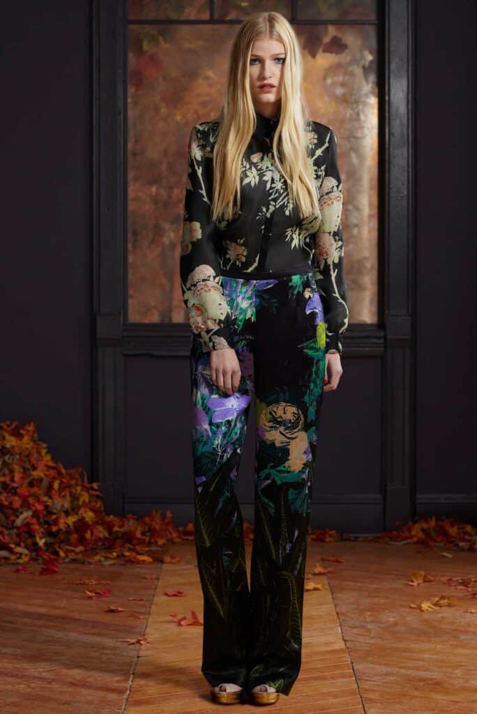 Woman in floral blouse and wide-leg pants stands indoors, with autumn leaves scattered on the floor.