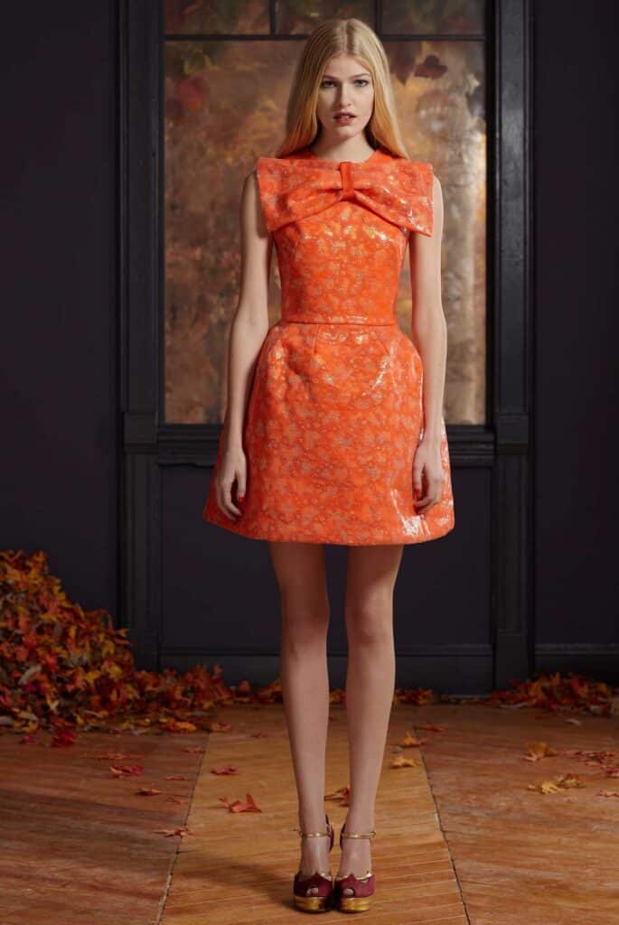 Woman in an orange dress with a large bow stands indoors, autumn leaves scattered on the floor.