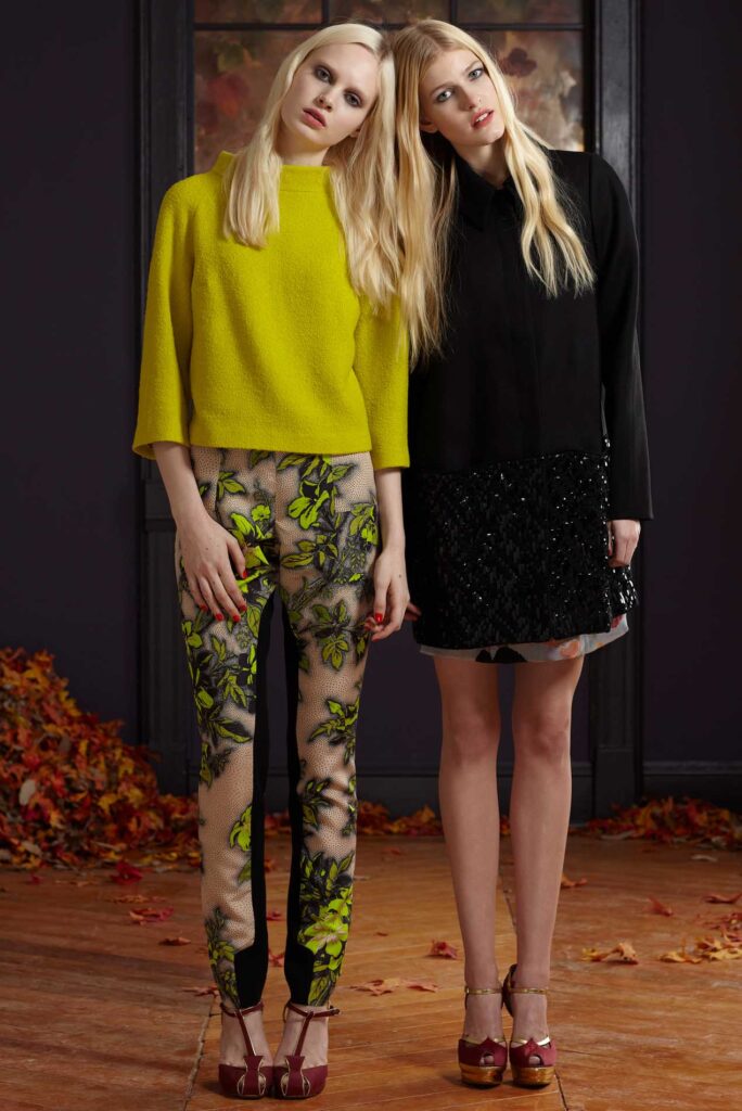 Two women pose indoors; one wears a yellow top and floral pants, the other a black dress. Leaves on the floor.