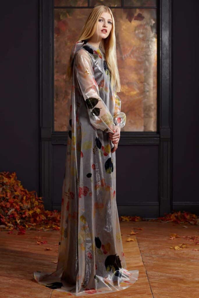 Woman in a long, sheer, floral-patterned dress stands indoors near scattered autumn leaves and a large window.
